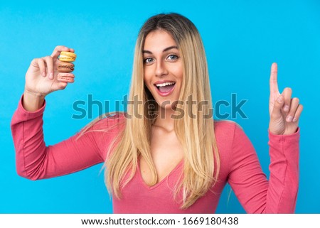 Young Uruguayan woman over isolated blue background holding colorful French macarons and pointing up a great idea