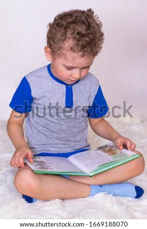 A little boy sits on a fluffy bedspread and reads a book. Teaching children to read, a photo