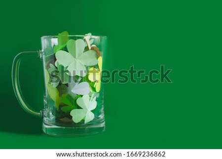 Bear transparent cup with different shades of green four-leafed paper shamrocks on green background. Saint Patricks day and lucky concept. Place for text