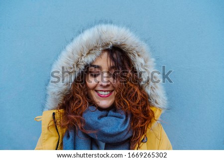 Caucasian woman looking down with a hood winter yellow coat. Portrait of beautiful female isolated on blue background with copy space. Bright complementary colors. Travel and winter holidays concept.