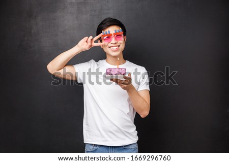 Birthday, celebration and party concept. Portrait of happy friendly smiling asian man enjoying b-day, wear funny glasses hold cake with lit candle, making wish, show peace sign near eye