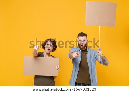 Angry protesting young people guy girl hold protest signs broadsheet blank placard on stick point fingers on camera isolated on yellow background. Protests strikes pickets concept. Youth against city