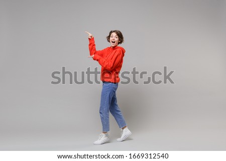 Side view of surprised young brunette woman girl in casual red hoodie blue jeans posing isolated on grey background in studio. People lifestyle concept. Mock up copy space. Point index fingers aside Royalty-Free Stock Photo #1669312540