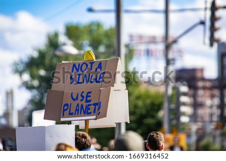 A small cardboard placard is seen up close, with blurry city buildings in background, saying not a single use planet, with copy space to right