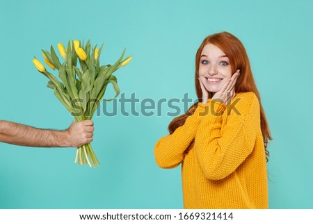 Excited young redhead woman girl in yellow sweater posing isolated on blue turquoise background in studio. People lifestyle concept. Mock up copy space. Taking bouquet of yellow tulips in male hand