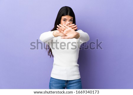 Young brunette woman over isolated purple background making stop gesture with her hand to stop an act