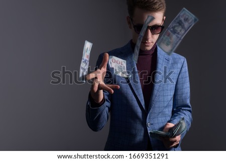 Portrait of a young guy in sunglasses and a business suit, with a serious look, scatters dollar bills. On a gray background.