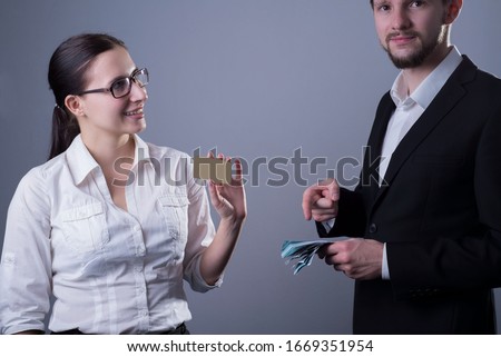 two young business people.A smiling girl is holding a plastic card in her hand, looking at the guy who is pointing at the camera while pointing to the press of cash dollar bills in his hand