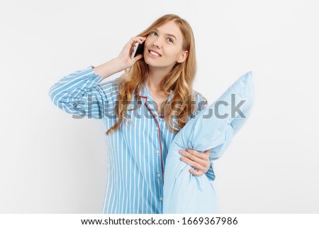 Happy attractive young girl in pajamas with pillow smiling talking on phone on white background