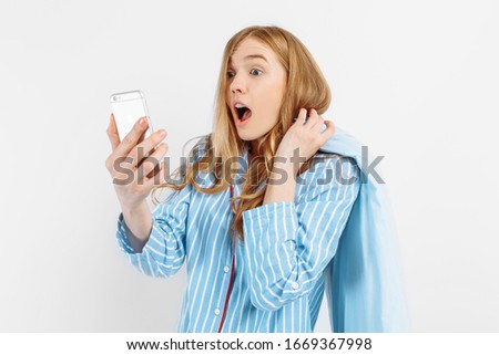Shocked surprised a young girl in pajamas with a pillow in shock looking at the screen of the smartphone on a white background