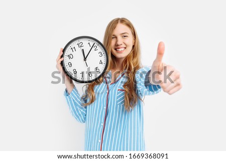 Happy girl wearing pajamas holding an alarm clock shows the gesture a class, waking early in the morning on a white background Royalty-Free Stock Photo #1669368091