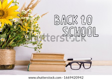 The inscription marker on white board, Back to School. A table with books, a bouquet of flowers, glasses and attributes for writing.