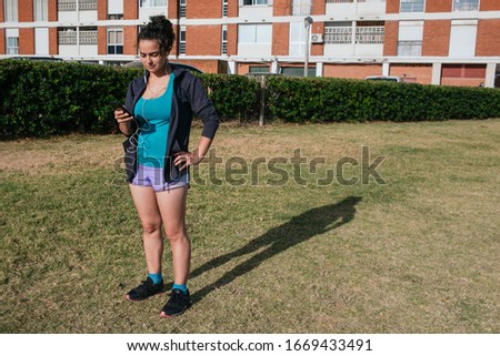 white, caucasian, latin woman, with shorts and sportswear, using her cell phone with headphones, listening music on the grass, before running, turning on a mobile app