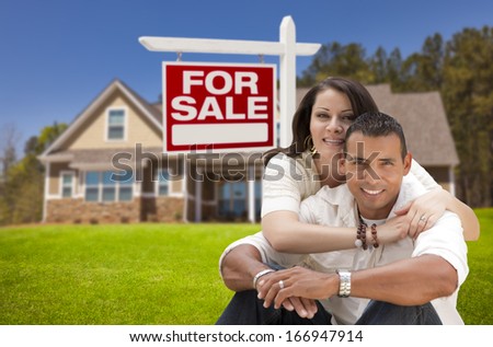 Young Happy Hispanic Young Couple in Front of Their New Home and For Sale Real Estate Sign.