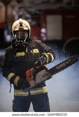 Firefighter portrait wearing full equipment, oxygen mask, and chain saw. Smoke and fire trucks in the background.
