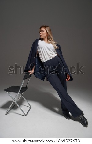 Stylish young woman in costume standing leaning on chair in studio at gray background. Fashion style portrait