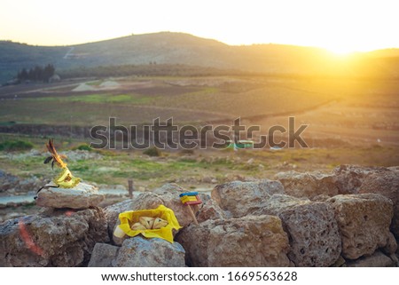 Golden carnival mask with feathers, a basket with triangle Purim cookies and a noise maker on the outdoor stone nature background with the skyline at sunset, Purim greeting postcard concept