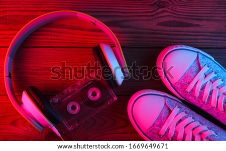 Stereo headphones with audio cassette and sneakers on wooden background. Neon red blue light. Gradient glow. Retro media. Top view