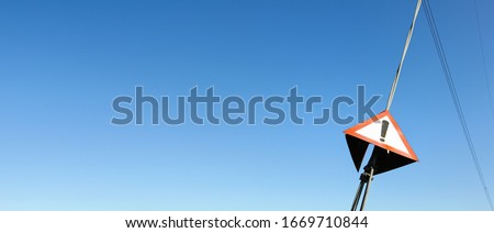 Red triangle with exclamation mark on steel cable of electric pylon, clear sky in background - wide photo, space for text left side
