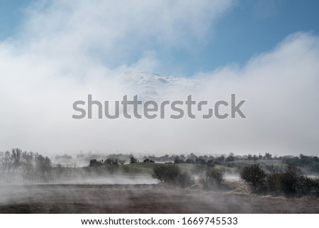 Lowland and fields covered with fog. Mountains, low clouds and snow, in the background.