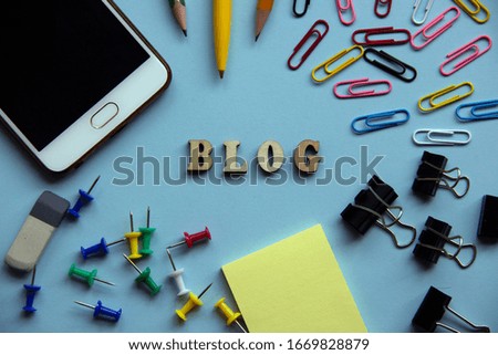 The word BLOG made of wooden letters, around a paper clip, pen, pencils, paper, binders, eraser, buttons, mobile phone. Creative concept. Top view, flat lay.