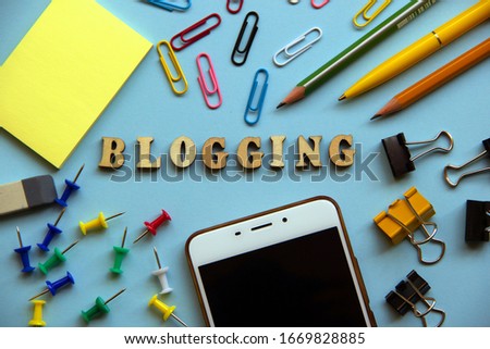 The word BLOGGING made of wooden letters, around a paper clip, pen, pencils, paper, binders, eraser, buttons, mobile phone. Creative concept. Top view, flat lay.