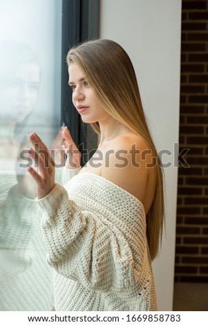 Beautiful young woman in sweater looking through the window