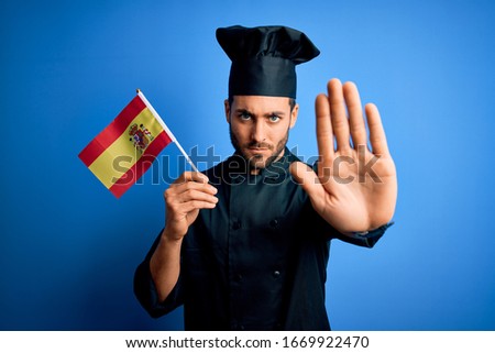 Young handsome cooker man with beard wearing uniform holding spain spanish flag with open hand doing stop sign with serious and confident expression, defense gesture
