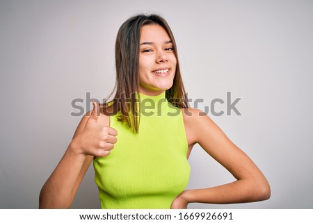 Young beautiful brunette girl wearing casual summer t-shirt over isolated white background doing happy thumbs up gesture with hand. Approving expression looking at the camera showing success.
