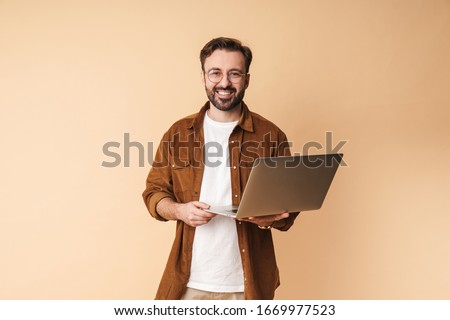 Image of a happy cheery optimistic young unshaved man isolated over beige wall background using laptop computer.
