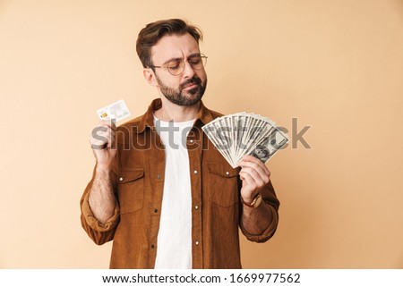 Photo of a serious young unshaved man isolated over beige wall background holding money and credit card.