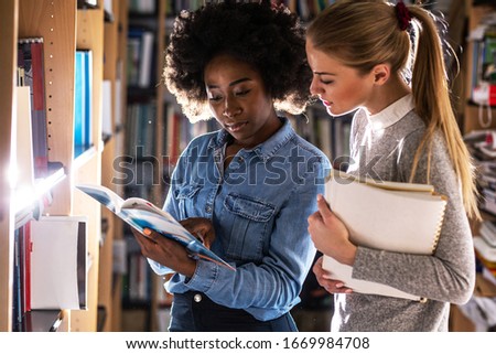 Two female college students reading  a book in campus library.	