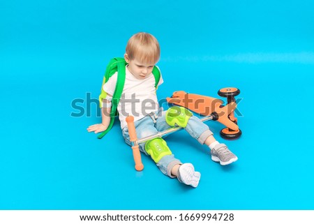 Kid blond boy in protection on his knees and elbows with a backpack fell from a scooter on a bright blue background. Studio shot concept for advertising security systems for active children sports.