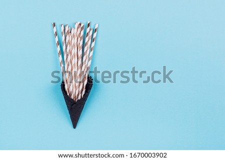 Cocktail straws with golden stripes in black ice-cream horn on pastel blue background with copy space. Creative and moody picture. Top view.