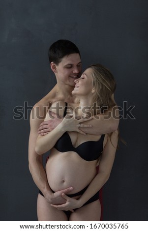 Happy man embracing his pregnant girlfriend and looking at camera. Loving husband gently hugging pregnant wife at home. Young couple expecting a baby.