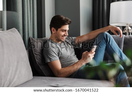 Portrait of an attractive smiling young bearded man wearing casual clothes sitting on a couch at the living room, using mobile phone Royalty-Free Stock Photo #1670041522