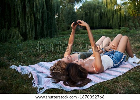 Two sisters, lying on blanket on grass in park at sunset, taking selfie for social media. Young pretty girls, wearing jeans shorts, green khaki beige tops, having fun outside. Leisure summertime.