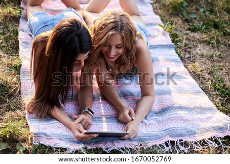 Two sisters, lying on blanket on grass in park in summer, watching fun videos on tablet. Pretty girls, wearing light blue jeans shorts and green beige top, relaxing outside, smiling, laughing.