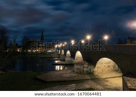 Historic illuminated stone bridge in Regensburg, Bavaria with cathedral in background at night
