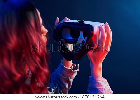 Rear view of girl with curly hair holding virtual reality glasses in red and blue neon in studio.