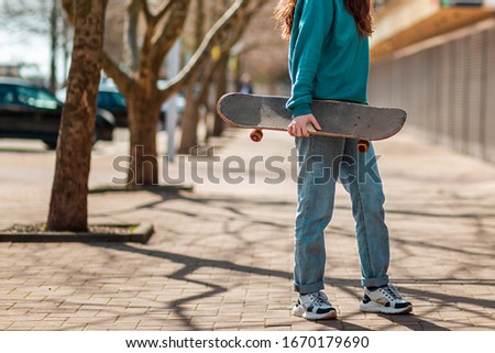 A young hipster teen walks down the street with a skateboard in her hands and turns back. In the background, an alley. Close up. Concept of sports lifestyle and street culture
