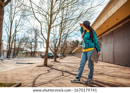 A young Caucasian hipster woman stands with a skateboard in her hand and looks into the distance. In the background, an alley. Concept of sports lifestyle and street culture