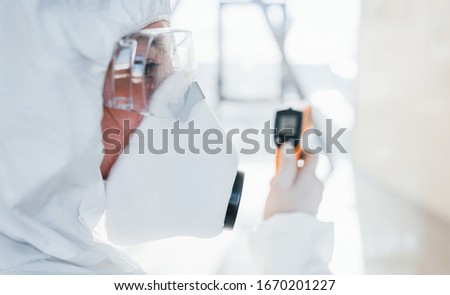 Female doctor scientist in lab coat, defensive eyewear and mask standing indoors with infrated thermometer.