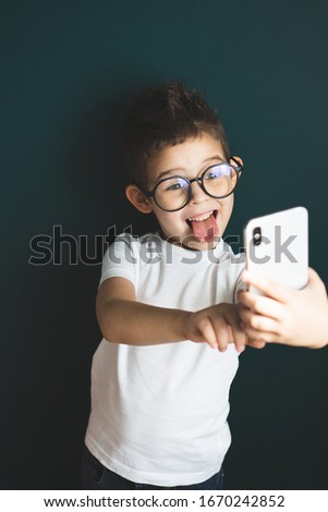 Caucasian little boy in a empty white mock up t-shirt use phone or making selfie on smartphone