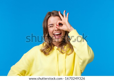 Close-up portrait of optimistic dreamy caucasian girl with blonde short hair, showing okay gesture over eye and wink happy, express positive enthusiastic emotion, standing blue background