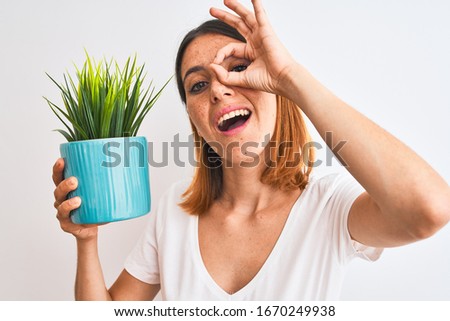 Beautiful redhead woman holding plant pot over isolated background with happy face smiling doing ok sign with hand on eye looking through fingers