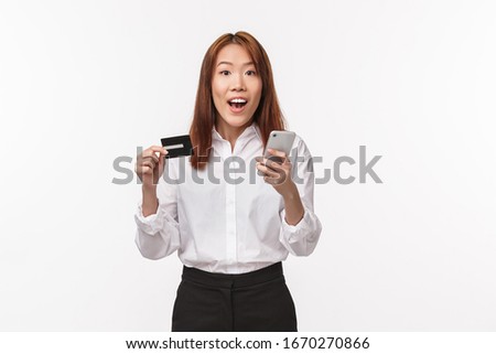Portrait of excited cute asian female in shirt and skirt, holding mobile phone and credit card, making internet order, shopping online, use bank deposit, register account in store, white background