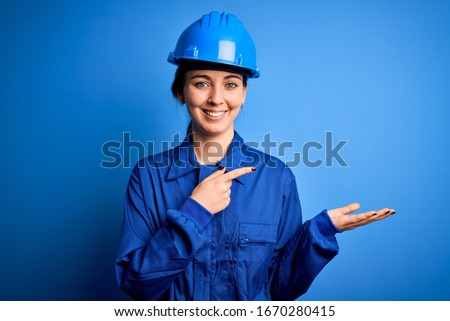 Young beautiful worker woman with blue eyes wearing security helmet and uniform amazed and smiling to the camera while presenting with hand and pointing with finger.