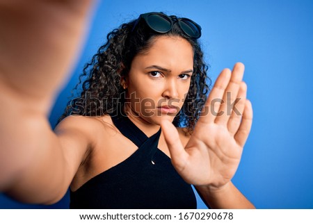 Young beautiful woman with curly hair wearing casual t-shirt making selfie by camera with open hand doing stop sign with serious and confident expression, defense gesture