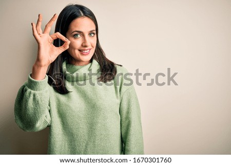 Young brunette woman with blue eyes wearing turtleneck sweater over white background smiling positive doing ok sign with hand and fingers. Successful expression.